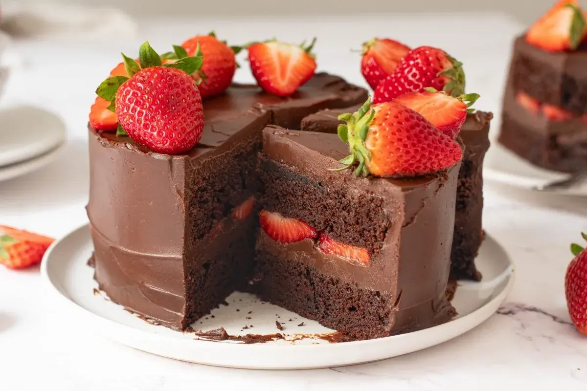 Deliciosa tarta de fresas y chocolate, cubierta de ganache y decorada con fresas frescas. Un trozo revela capas de bizcocho de chocolate y fresas.