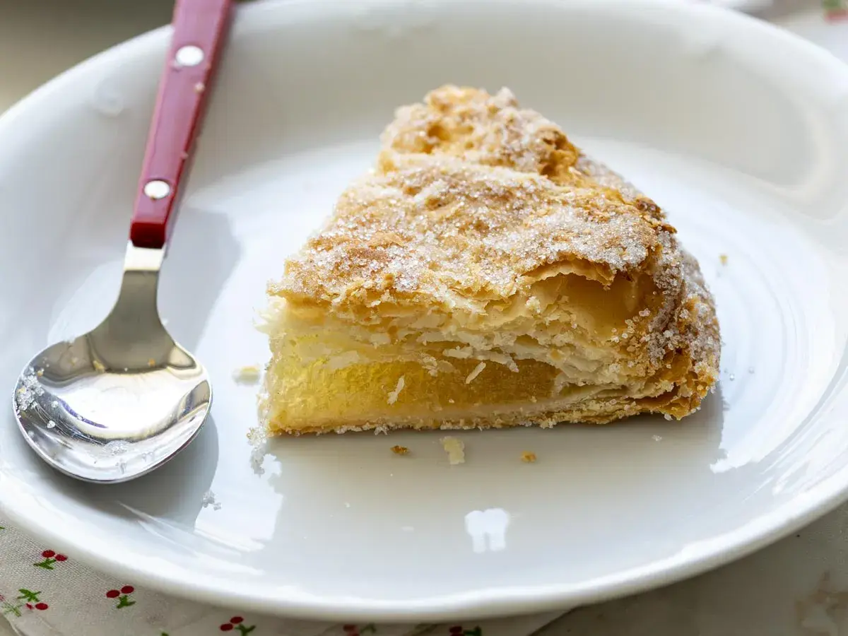 Un trozo de pastel cordobés, hojaldrado y cubierto de azúcar, espera ser disfrutado junto a una cuchara en un plato blanco.