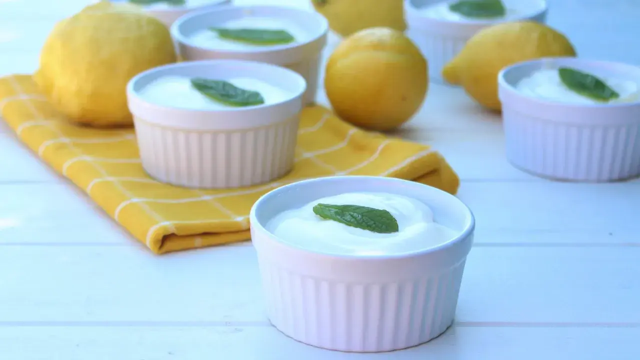 Delicioso mousse de limón mambo en ramequines blancos, adornado con hojas de menta. Limones frescos y una servilleta amarilla completan la escena.