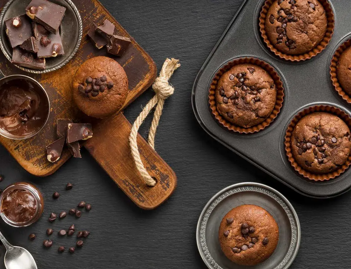 Deliciosos muffins de chocolate recién horneados, algunos en molde y otros listos para disfrutar, acompañados de trozos de chocolate y crema.