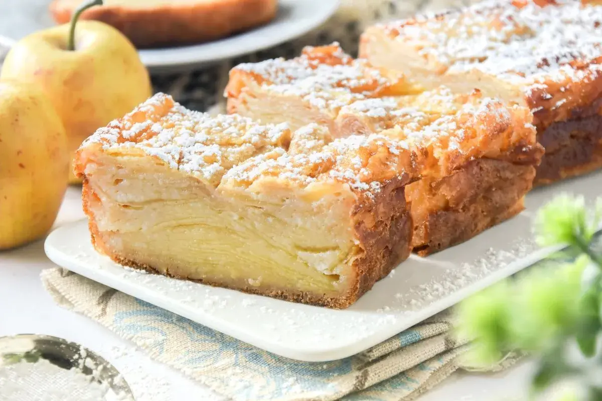 Porción de tarta de manzana con capas finas, espolvoreada con azúcar glas. Dos manzanas amarillas al lado.