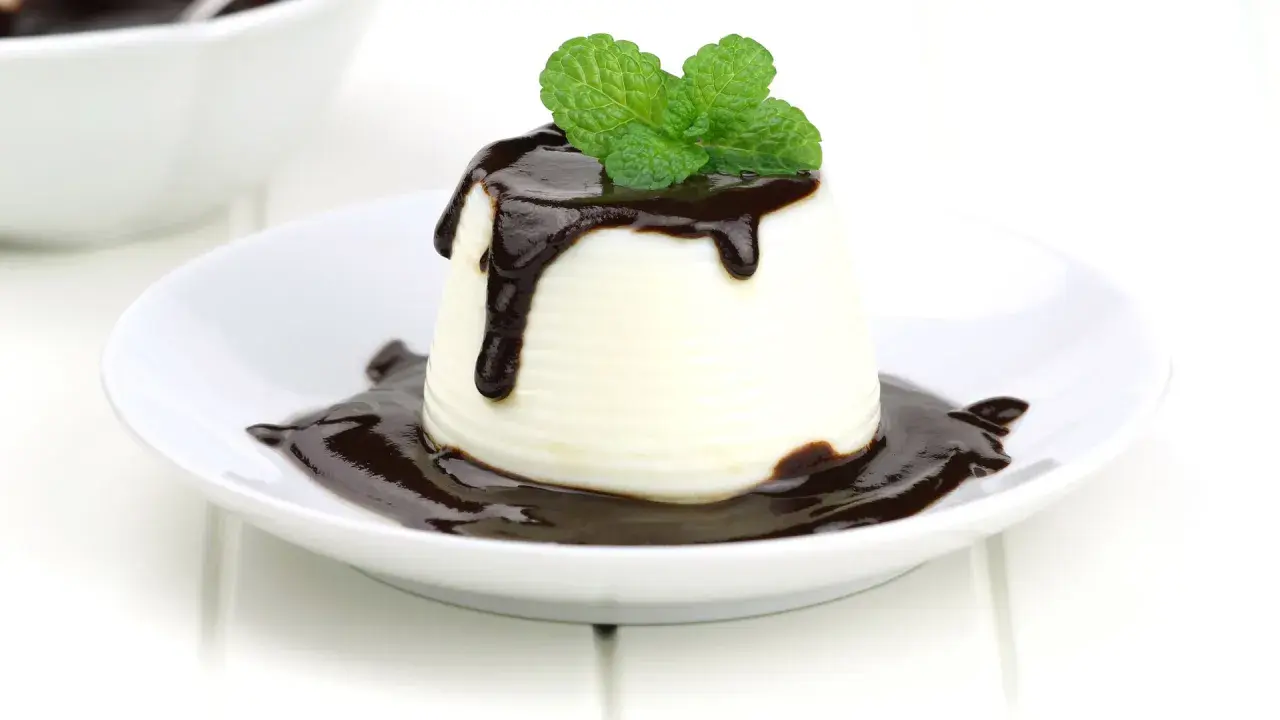 Deliciosa panna cotta de chocolate blanco, cubierta con salsa de chocolate y decorada con hojas de menta fresca.