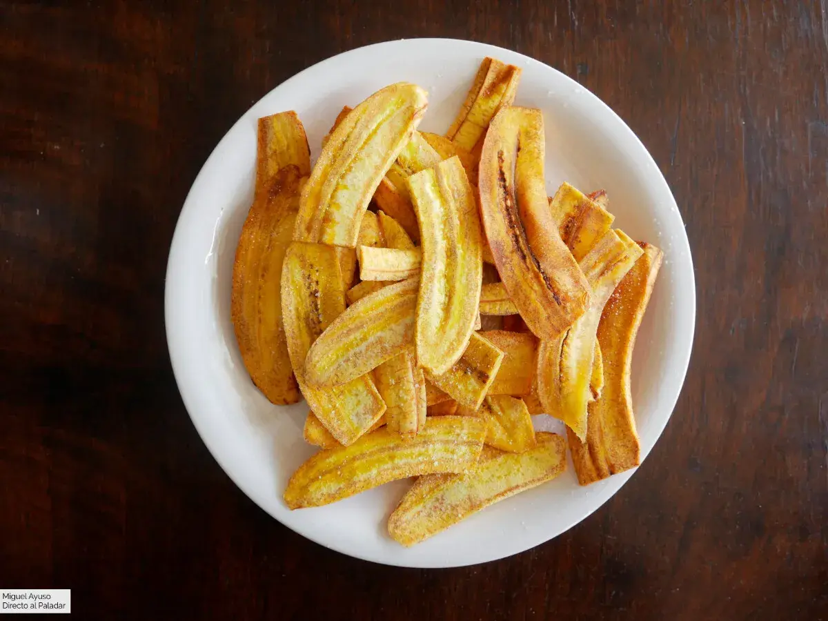 Un plato blanco lleno de crujientes rodajas de plátano frito, espolvoreadas con azúcar y canela.