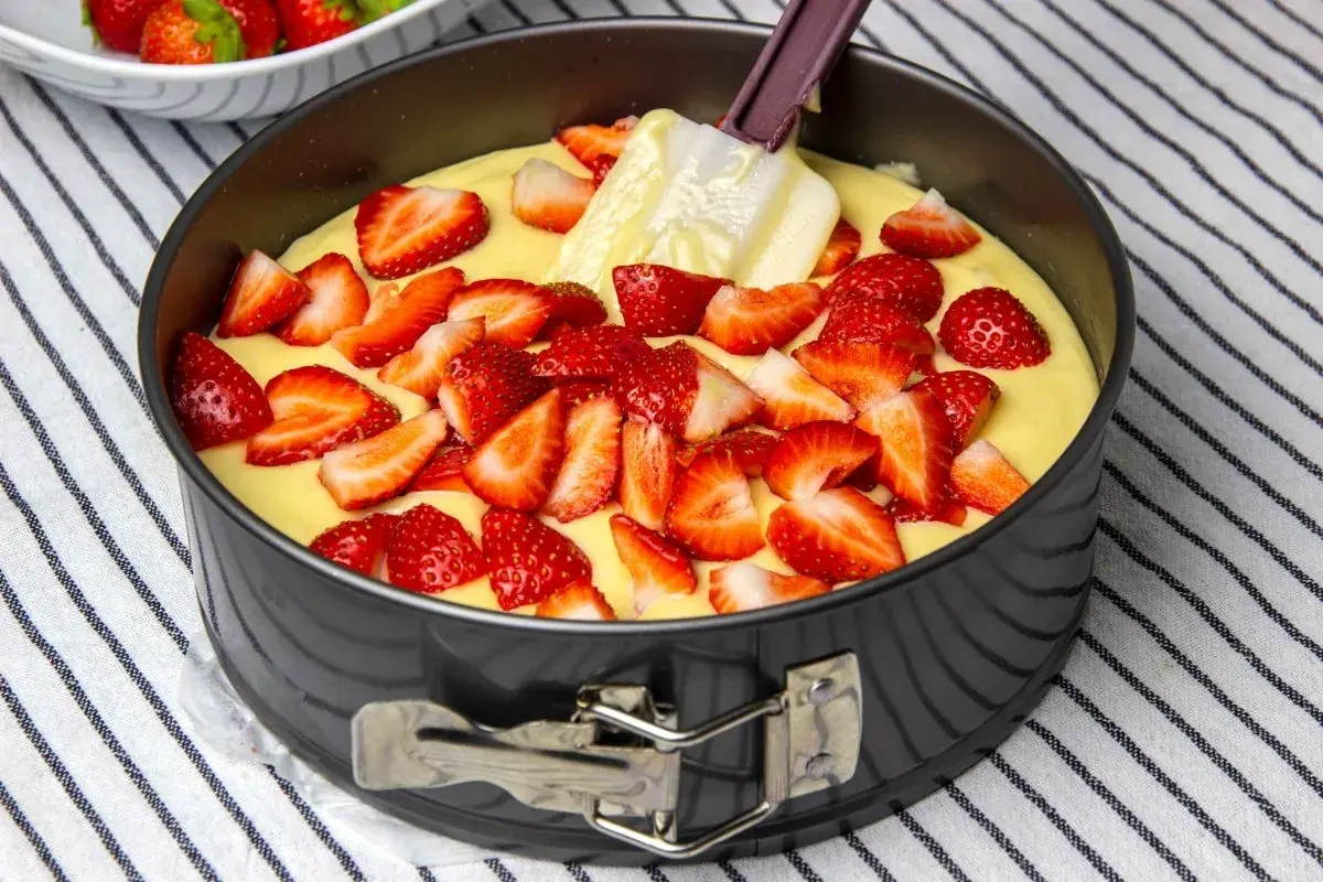 Bizcocho de yogur de fresa en molde, con fresas cortadas esparcidas sobre la masa y una espátula.