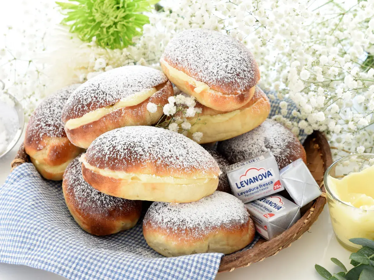 Bollos de mantequilla cubiertos de azúcar glas, rellenos de crema, junto a levadura y flores blancas.
