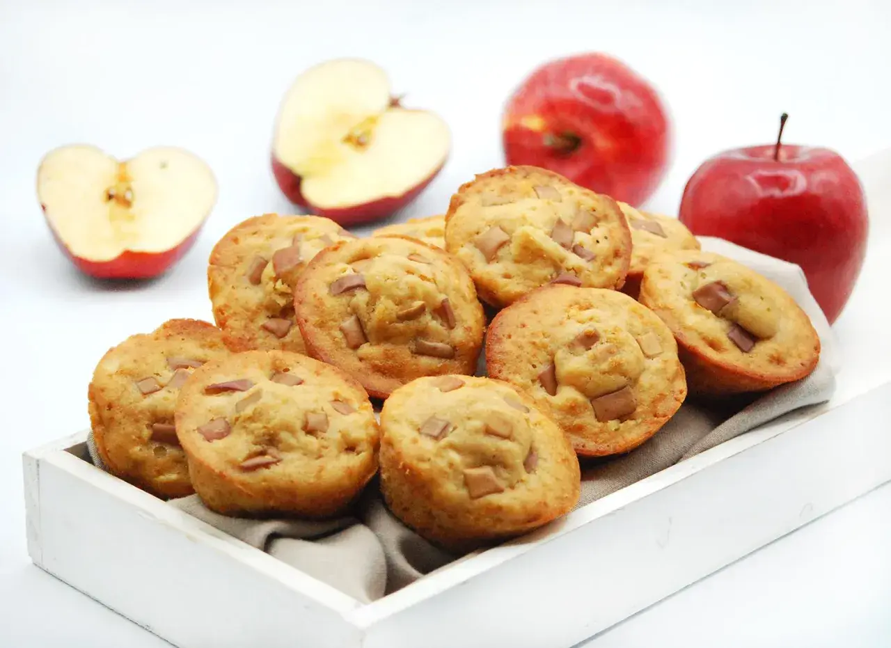 Deliciosas magdalenas de manzana, recién horneadas y listas para disfrutar. Dos manzanas rojas enteras y dos cortadas a la mitad acompañan esta tentación.