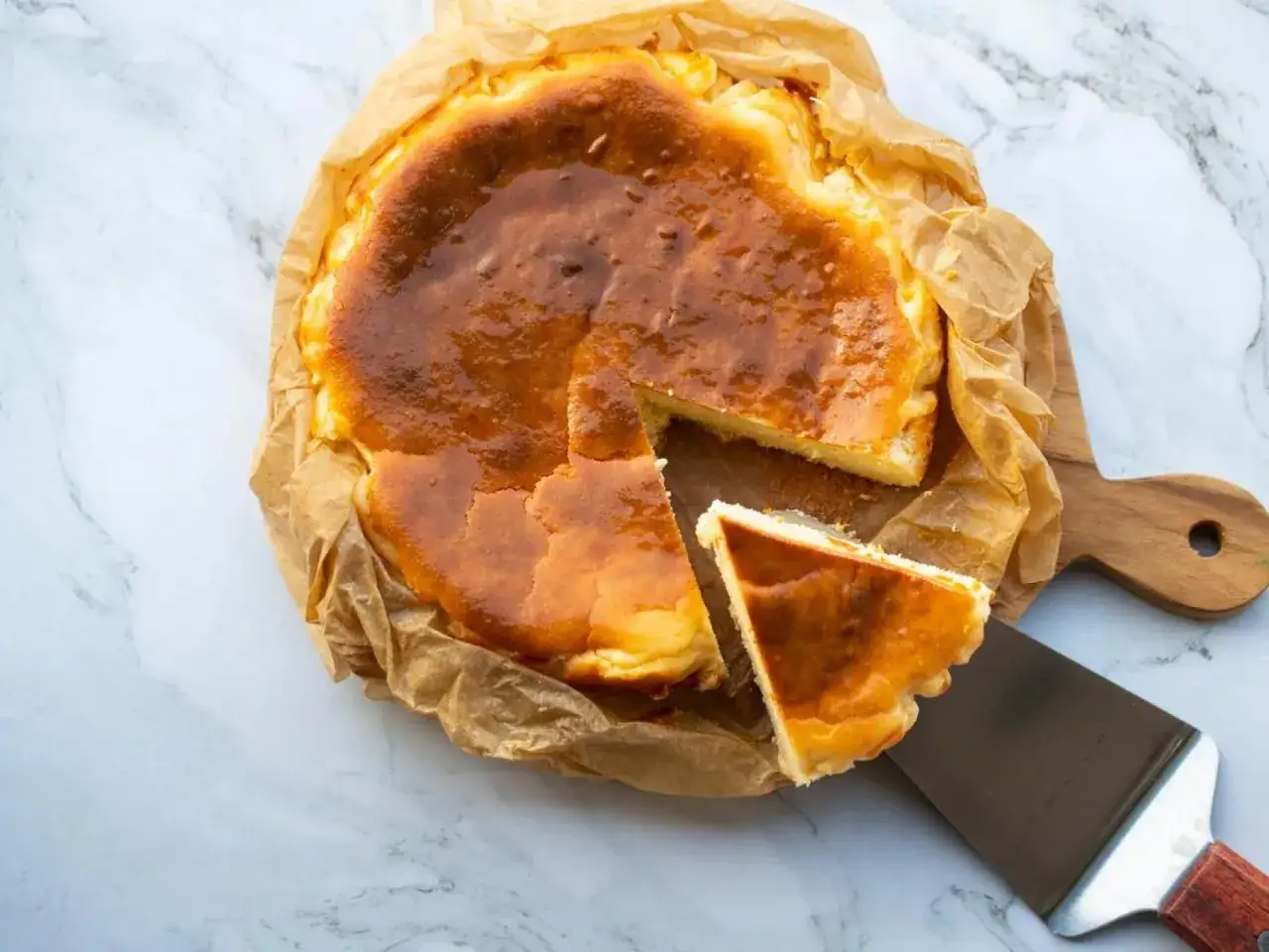 Tarta de queso congelada, dorada y cremosa, servida sobre papel de horno y tabla de madera. Una porción se sirve con una espátula.