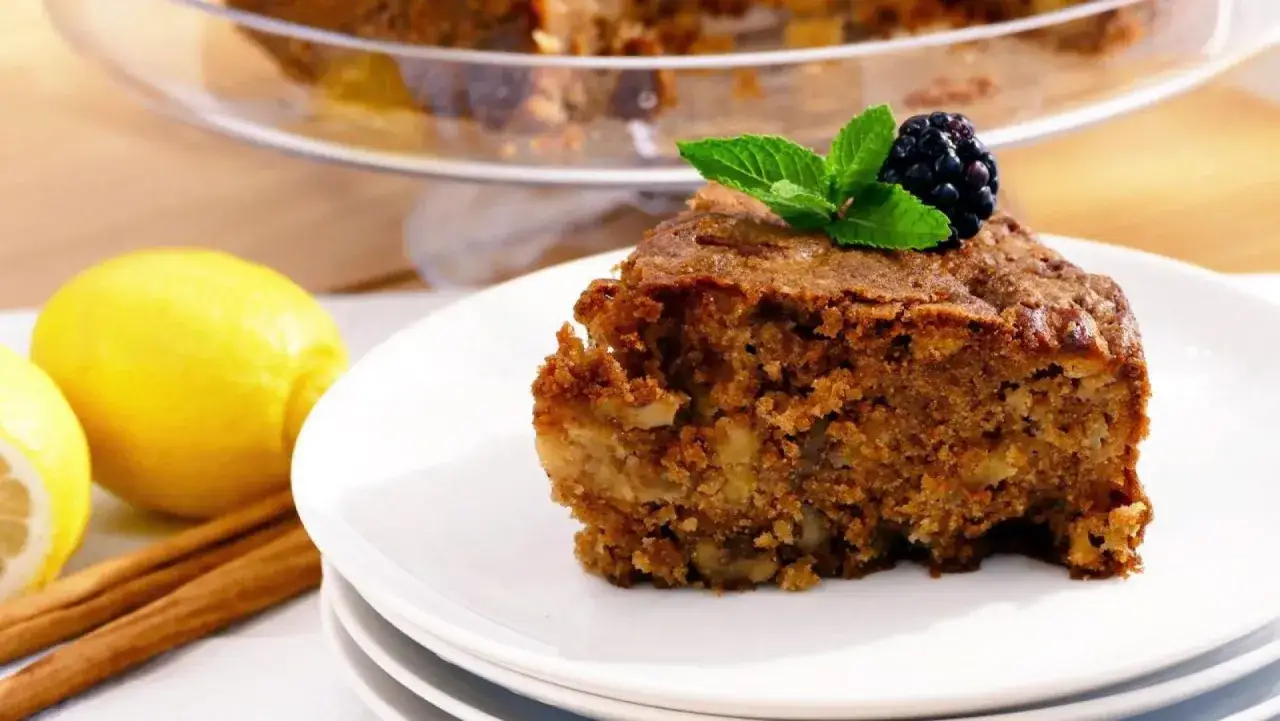 Un trozo de bizcocho de manzana y nueces, decorado con una mora y menta, junto a limones y canela.