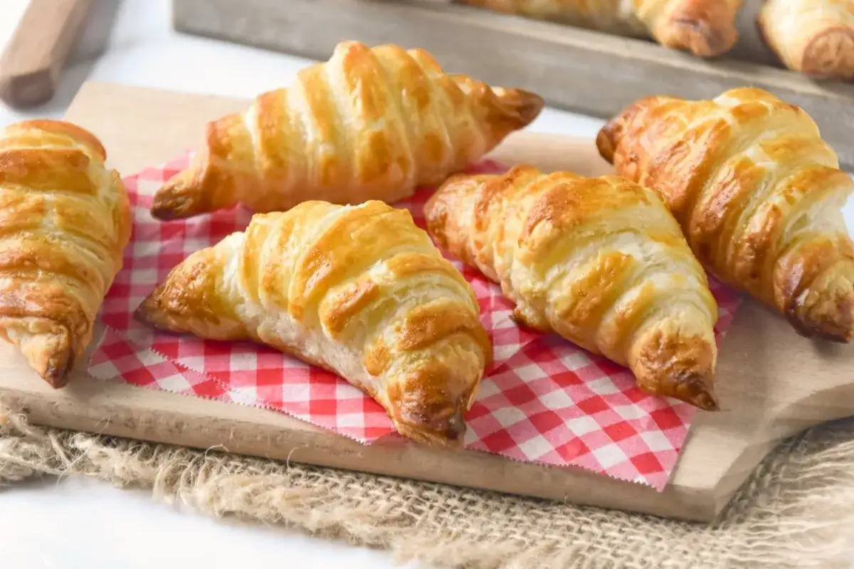 Seis croissants caseros dorados y hojaldrados sobre una tabla de madera y un mantel de cuadros rojos.