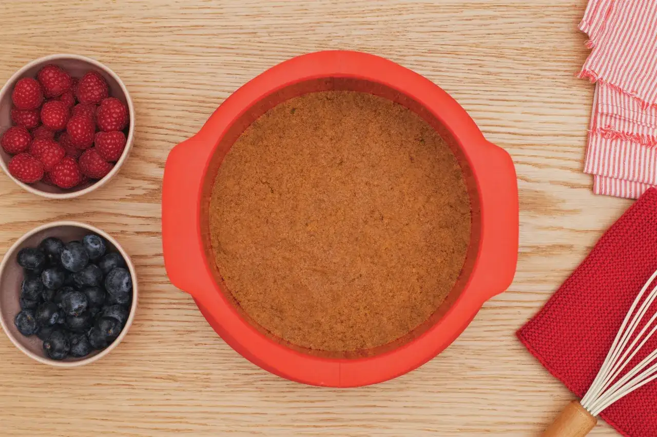Base de galleta lista en molde rojo para tarta de queso sin horno. Frambuesas y arándanos en cuencos.
