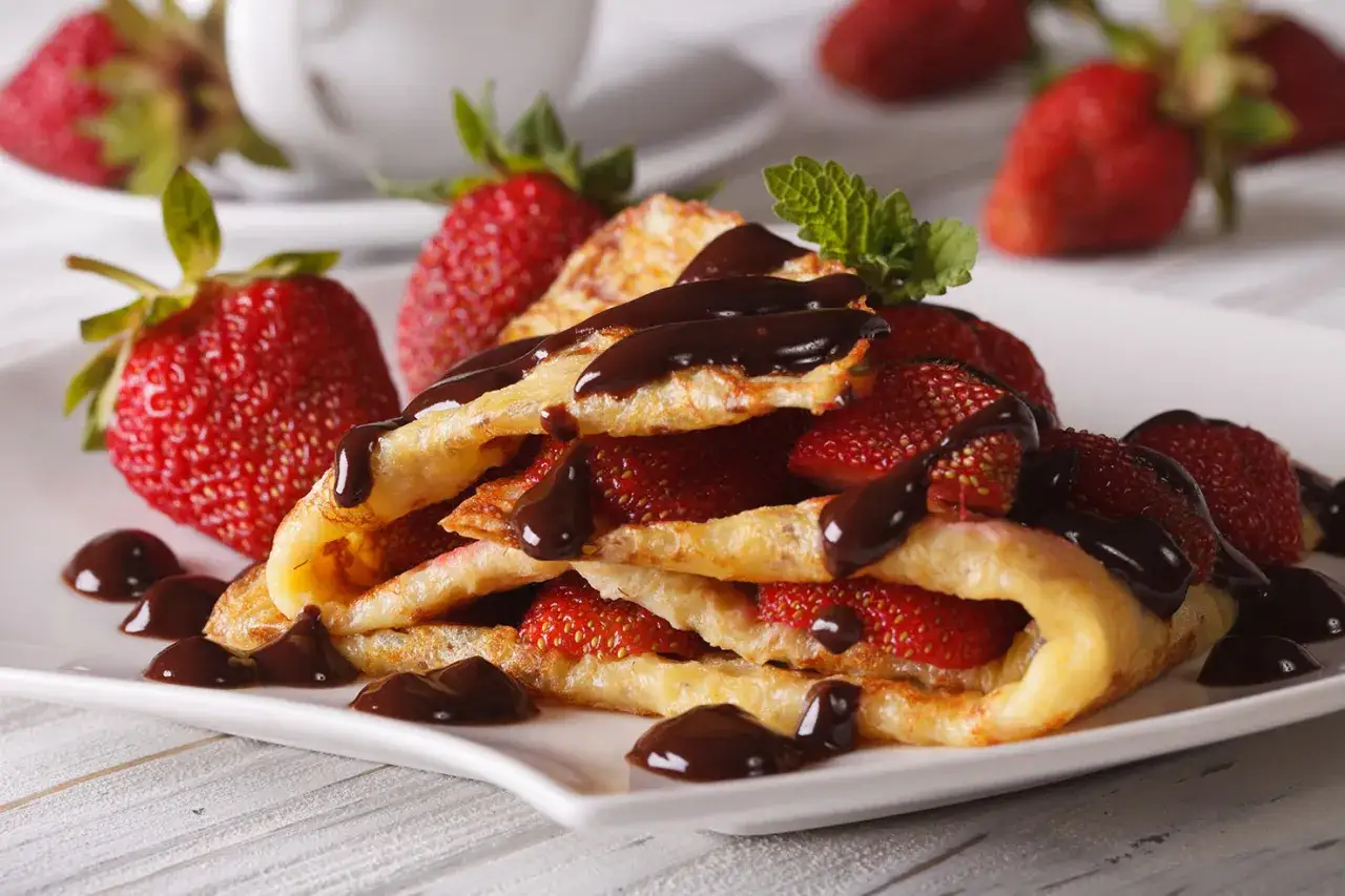 Deliciosos crepes de chocolate rellenos de fresas frescas, cubiertos con más chocolate derretido y adornados con una hoja de menta.