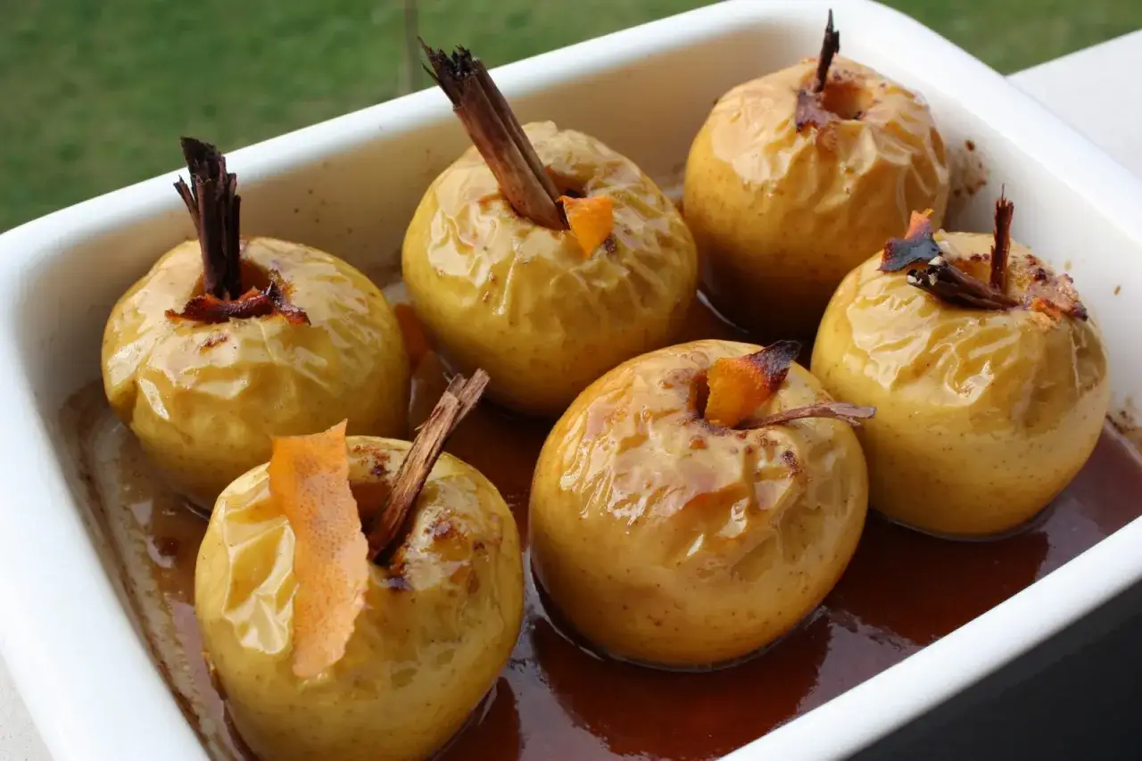Seis manzanas al horno doradas y brillantes, con canela y piel de naranja, reposan en una bandeja blanca con un sirope oscuro.