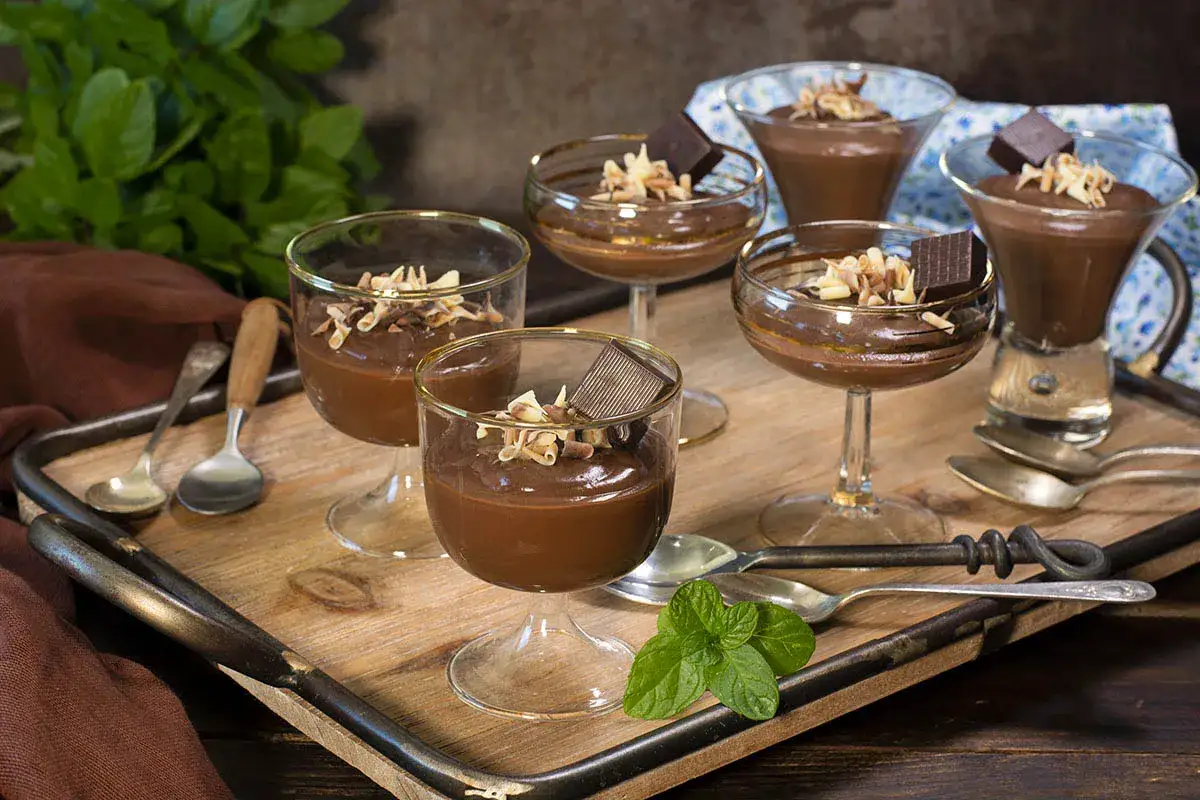 Deliciosas natillas de chocolate, cremosas y cubiertas con virutas de chocolate y un trozo de tableta. Servidas en copas, listas para disfrutar.