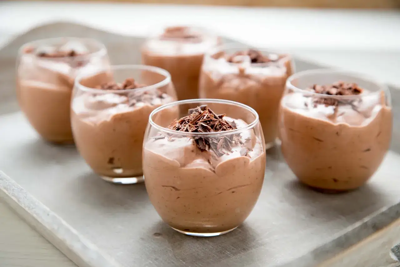 Seis copas de cremoso de chocolate, adornadas con virutas de chocolate, dispuestas en una bandeja de madera.