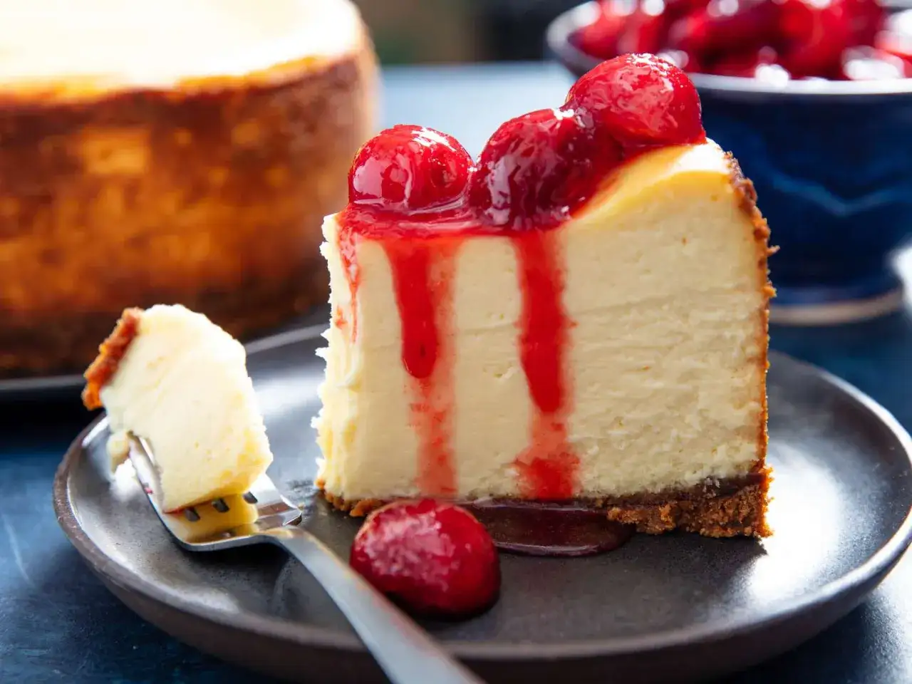 Un trozo de cremoso new york cheesecake cubierto con salsa de fresa y fresas frescas. Otro trozo en un tenedor.