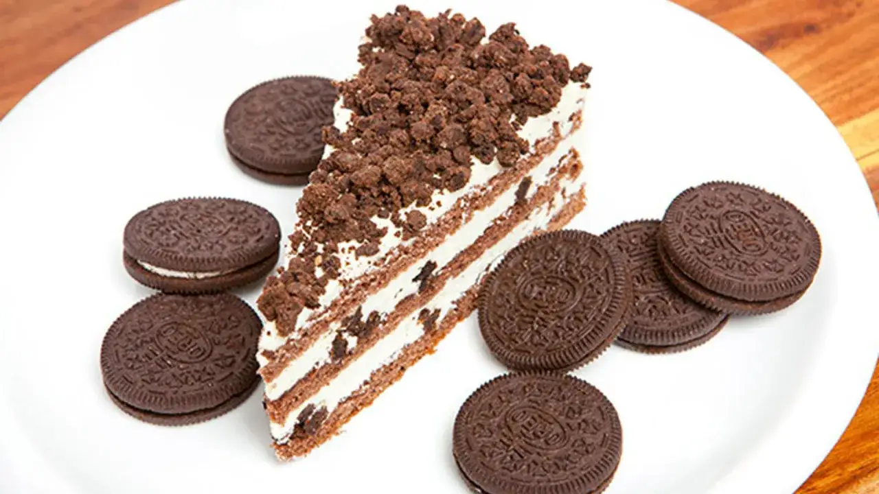 Rebanada de bizcocho de oreo con crema y trozos de galleta, rodeada de galletas oreo enteras en un plato blanco.