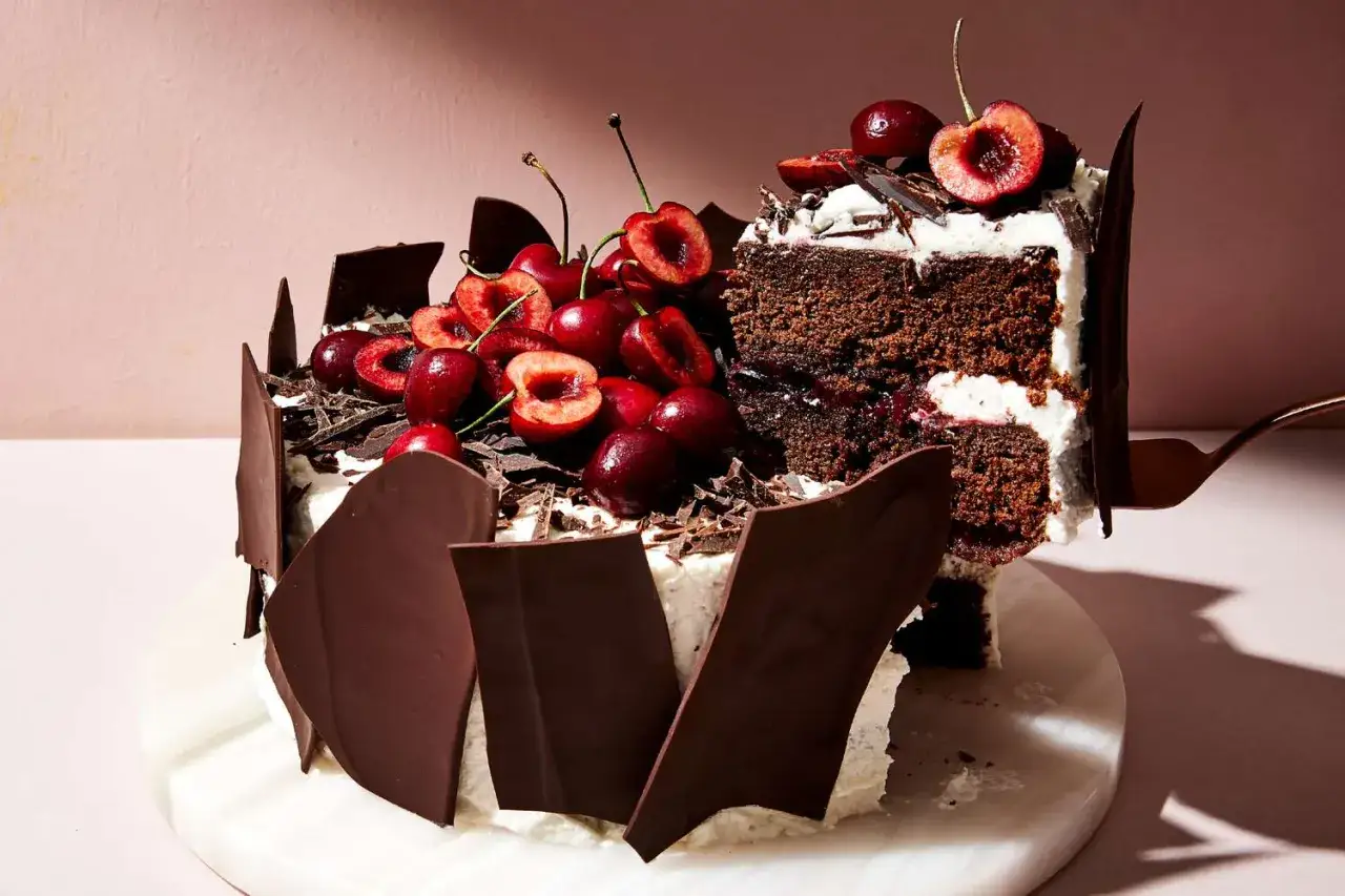 Tarta Selva Negra decorada con cerezas frescas, virutas de chocolate y trozos de chocolate negro. Una porci&oacute;n se sirve con un tenedor.