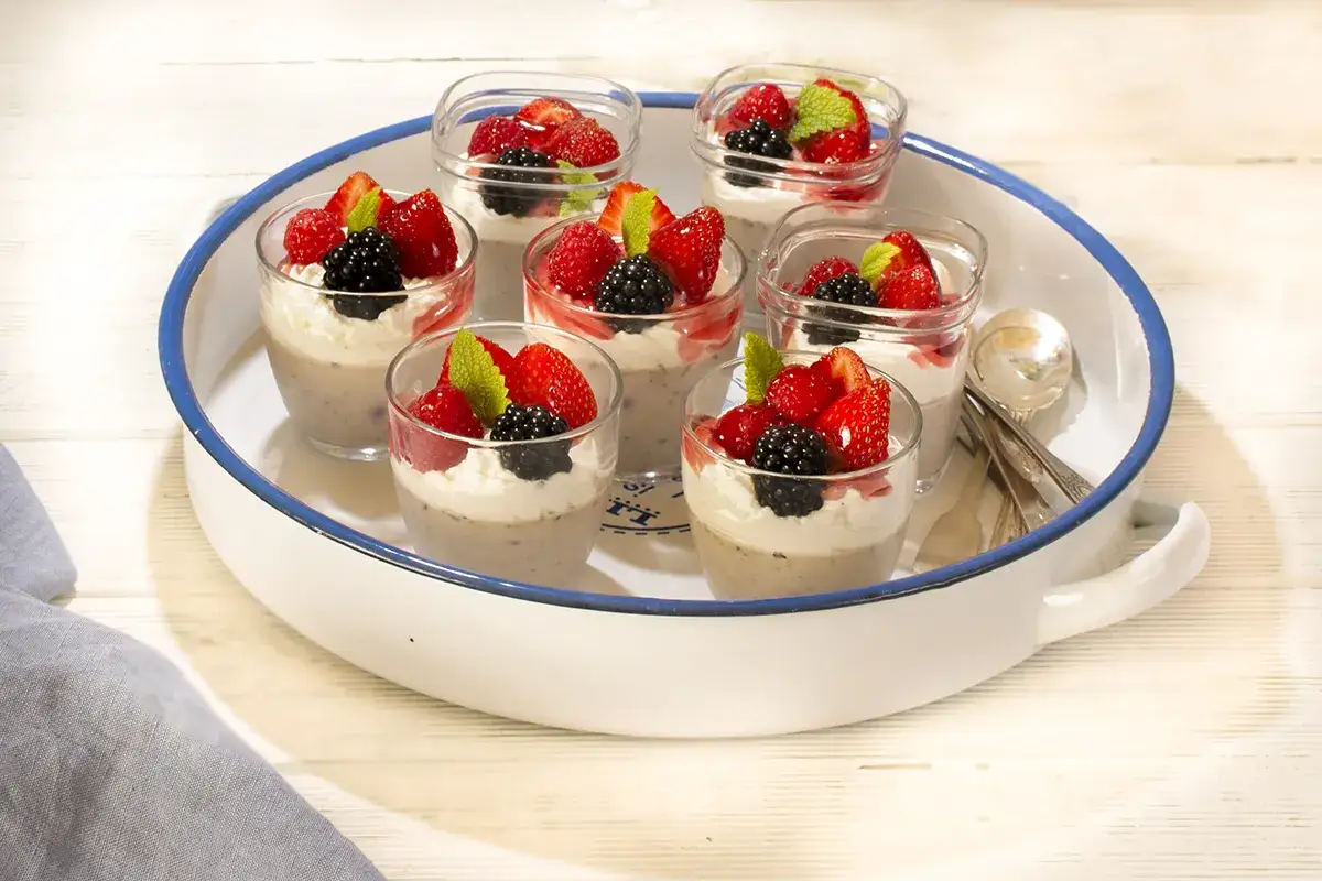 Seis copas de panna cotta de chocolate, decoradas con fresas, moras, frambuesas y menta, dispuestas en una bandeja blanca con borde azul.