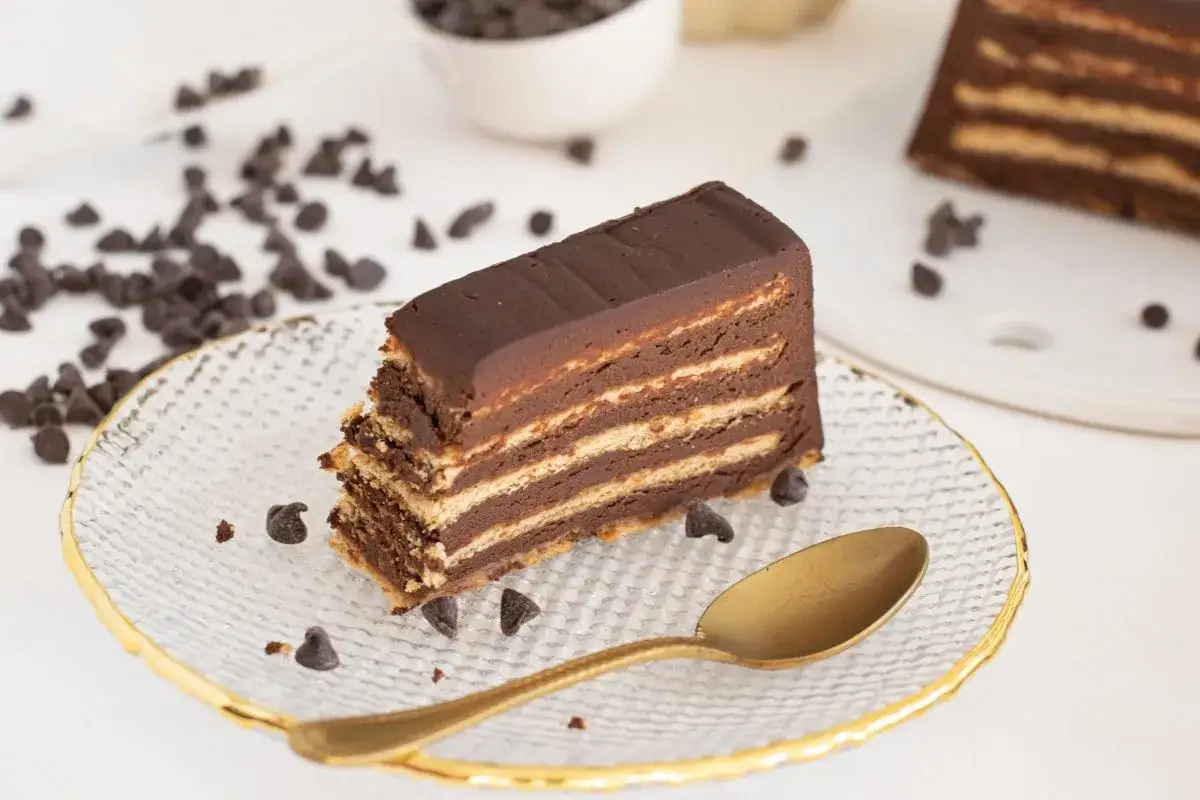 Rebanada de tarta de galletas y chocolate, con capas de galleta y crema de chocolate, decorada con chispas de chocolate y una cuchara dorada.