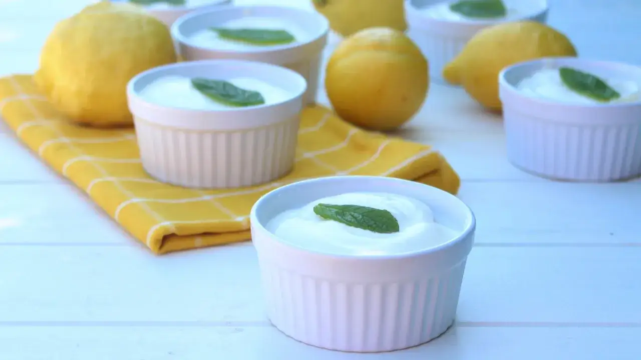 Delicioso mousse de lim&oacute;n mambo en ramequines blancos, adornado con hojas de menta fresca. Limones amarillos y un pa&ntilde;o a cuadros completan la escena.