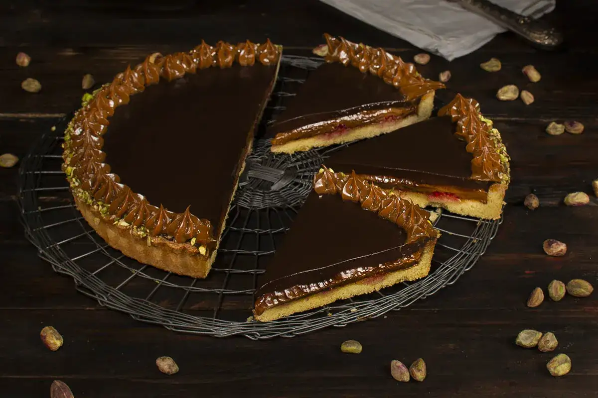 Tarta de dulce de leche con cobertura de chocolate brillante y bordes decorados con crema de dulce de leche y pistachos.