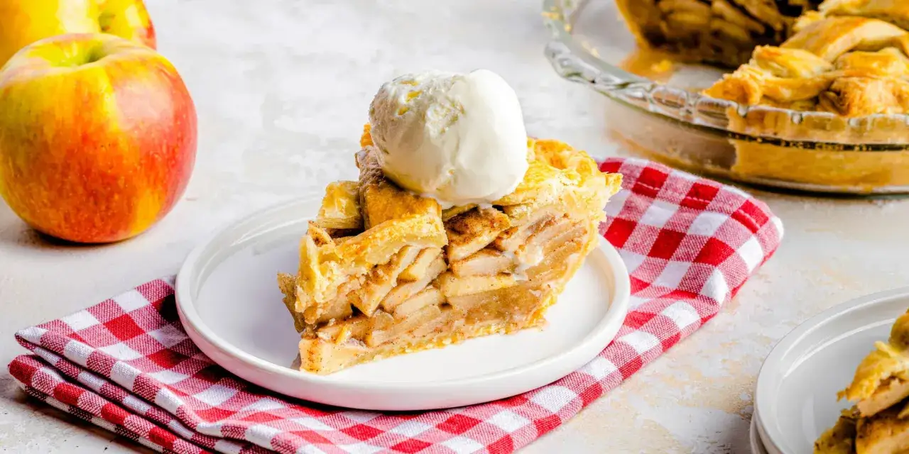 Rebanada de tarta de manzana casera con helado de vainilla, servida sobre un plato blanco con una servilleta a cuadros rojos.