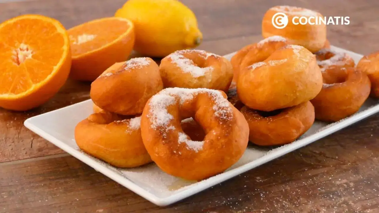 Rosquillas de naranja reci&eacute;n hechas, espolvoreadas con az&uacute;car, junto a naranjas y un lim&oacute;n.
