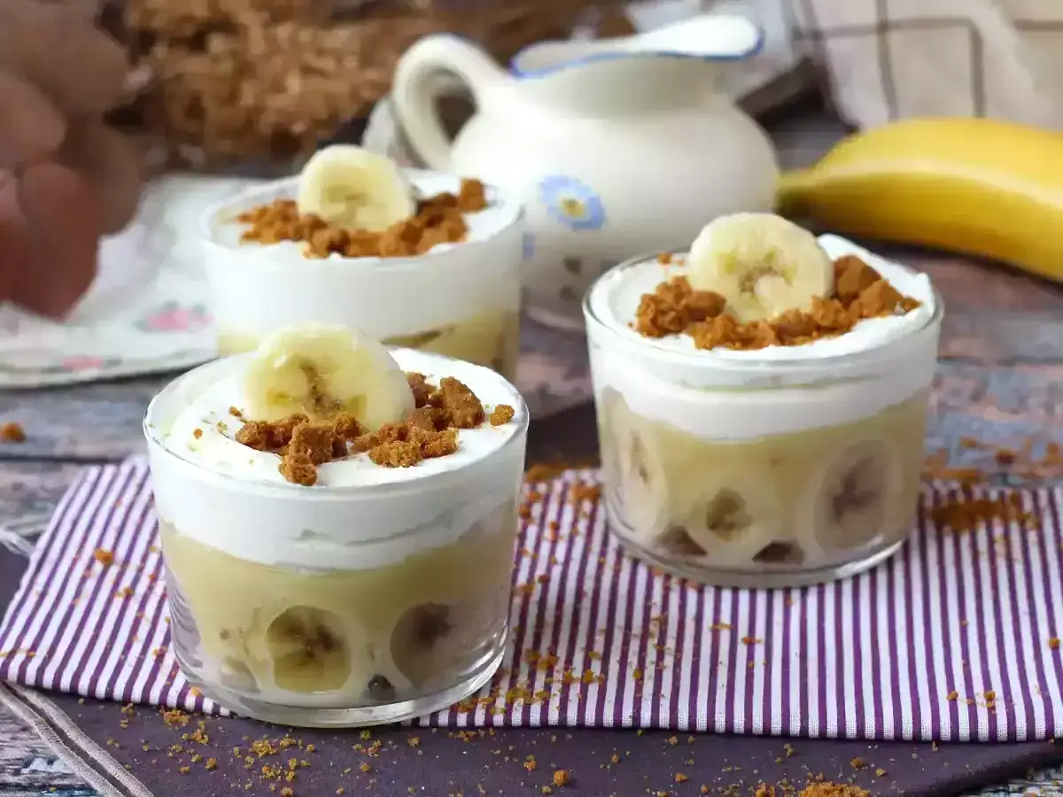 Delicioso pudin de platano en vasitos, coronado con crema batida, galleta triturada y una rodaja de platano.