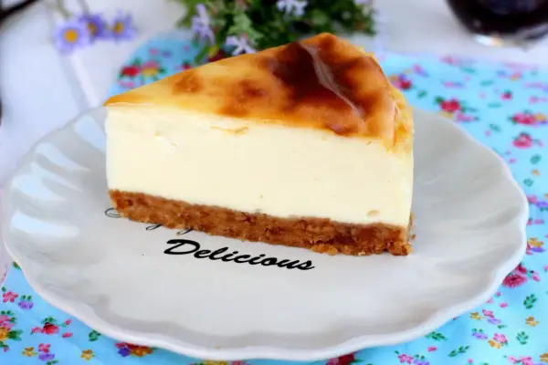 Rebanada de new york cheesecake con base crujiente y relleno cremoso, servida en plato blanco con la palabra "Delicious".